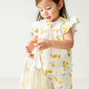 A toddler in a Makemake Organics Organic Linen Flutter Top and Shorts in Citron holds an empty string bag, looking curiously at her fingers. The background is a plain, light-colored wall.