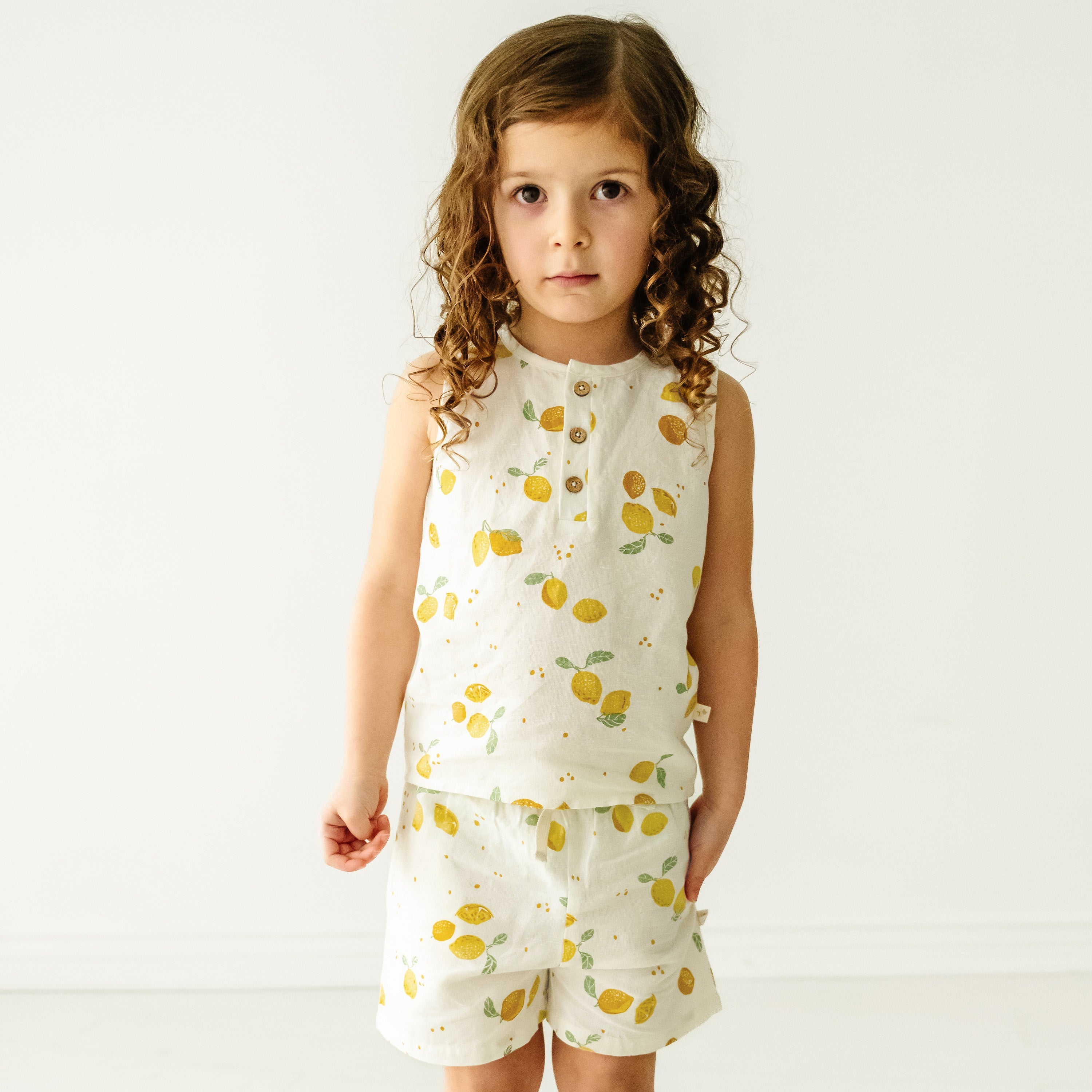 A young girl with curly hair stands facing the camera, wearing a white sleeveless Makemake Organics tank top and shorts decorated with yellow fruit prints, against a plain white background.