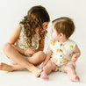 Two young children, one sitting and one toddler, interact lovingly. The older boy, with curly hair, smiles at the younger baby who sits upright. Both wear light outfits with lemon prints on a Makemake Organics Organic Linen Tank and Shorts Set in Citron.