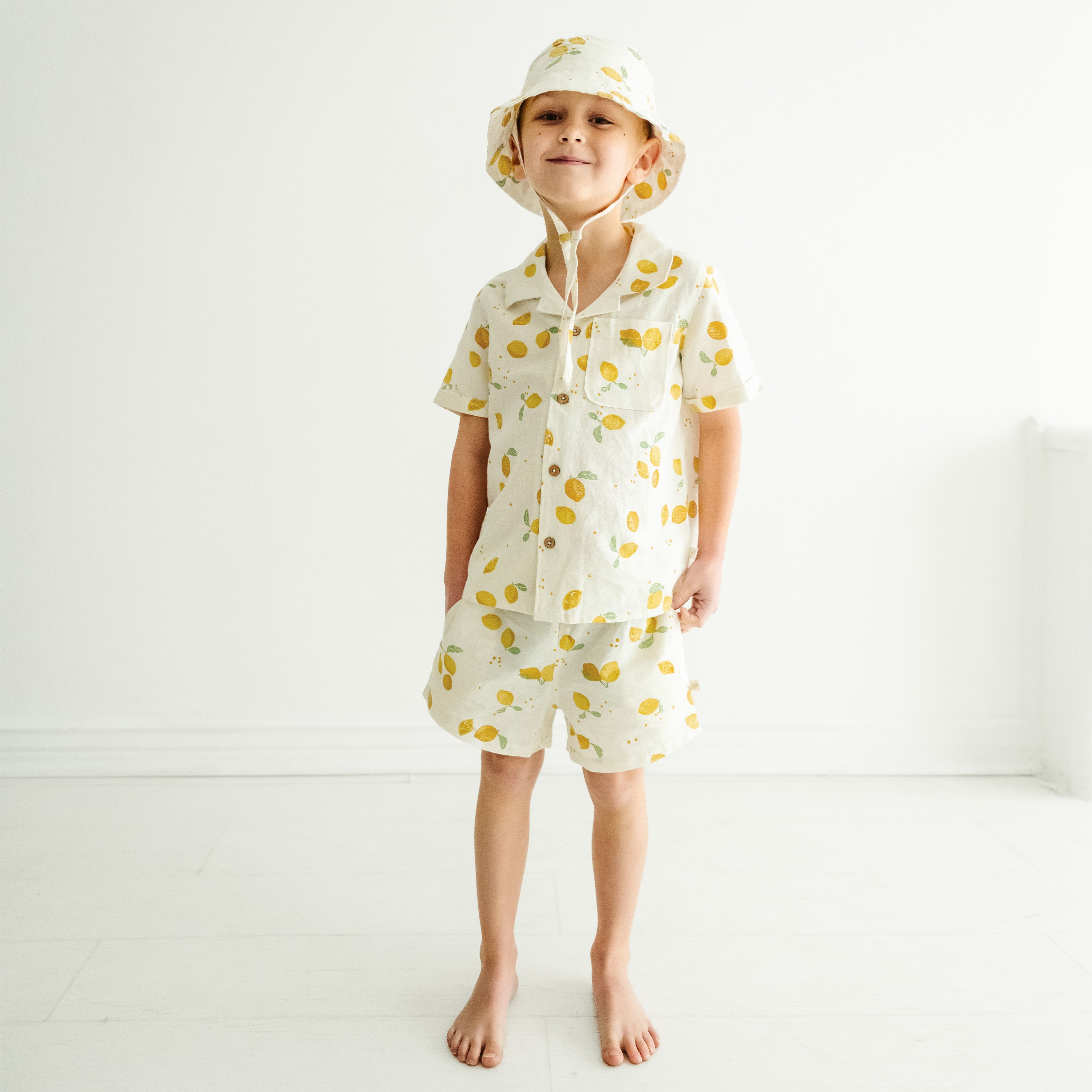 A young toddler stands barefoot in a light, airy room, wearing a Makemake Organics Organic Linen Shirt and Shorts Set adorned with lemon prints. The child looks directly at the camera with a subtle smile.