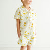 A young toddler wearing a summer outfit with a lemon print, Makemake Organics Organic Linen Shirt and Shorts Set - Citron, smiling happily, cropped view showing from the neck down against a clean white background.