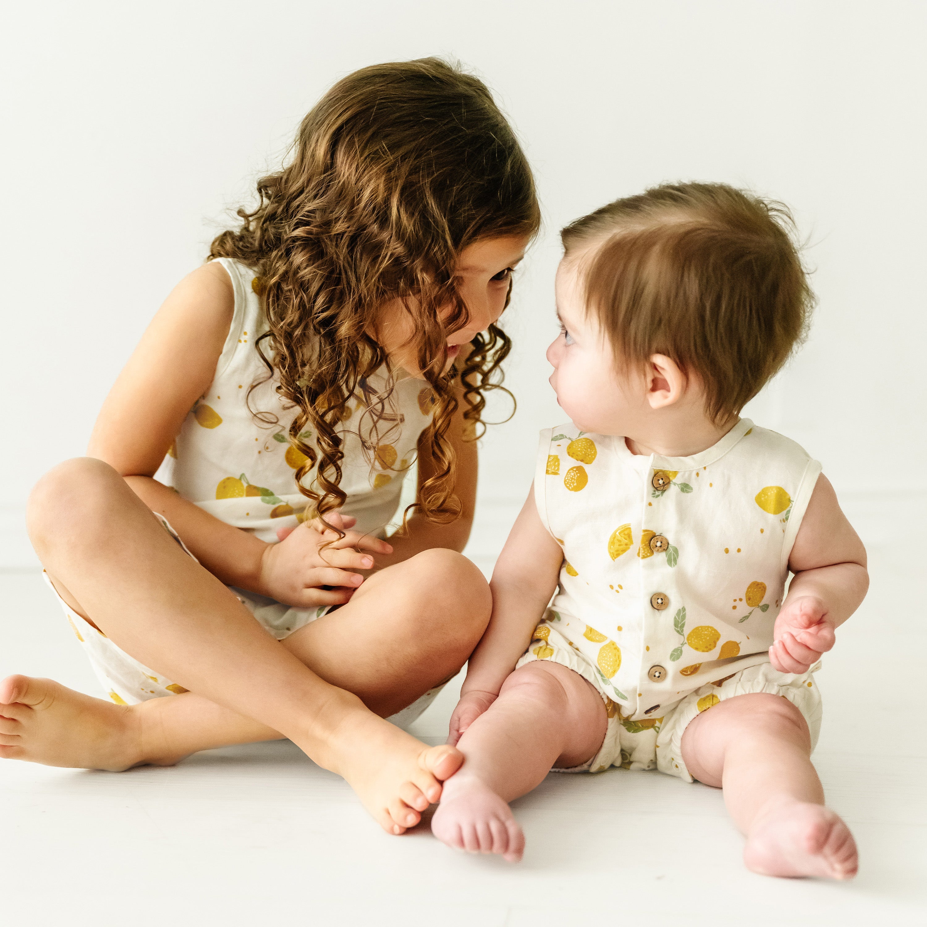 A young girl with curly hair sits cross-legged looking at a baby boy in a Citron Organic Linen Sleeveless Bubble Romper from Makemake Organics, who gazes back, both on a white background.