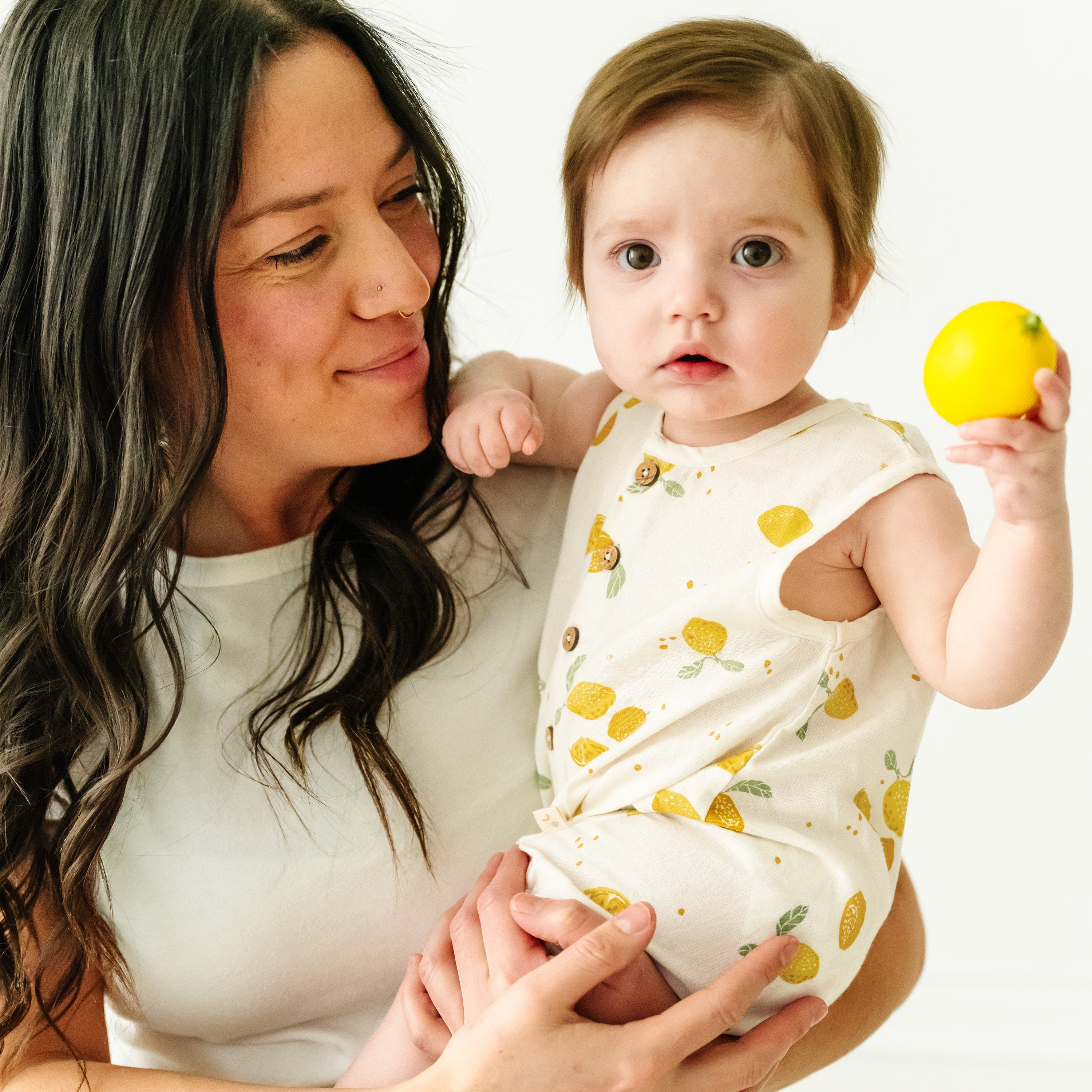A mother lovingly looks at her baby boy, who is holding a yellow ball. Both are on a white background, with the baby wearing a Makemake Organics Organic Linen Sleeveless Bubble Romper in Citron print.