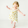 A joyful toddler with light hair, wearing a Makemake Organics Organic Linen Tiered Strap Dress in Citron with yellow floral patterns, playfully dancing barefoot in a bright room.