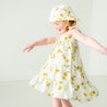 A young boy in a Makemake Organics Organic Linen Tiered Strap Dress - Citron and matching hat twirls joyfully in a bright, airy room.