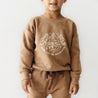 A young child wearing a cozy brown Organic Graphic Sweatshirt - Adventure from Organic Baby with the words "adventure" and "made for the mountains" printed on the front, standing against a