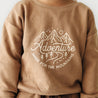 Close-up of a person wearing a brown Organic Graphic Sweatshirt - Adventure by Organic Baby, with a white graphic design depicting mountains and the words "adventure made for the mountains" printed on the front.