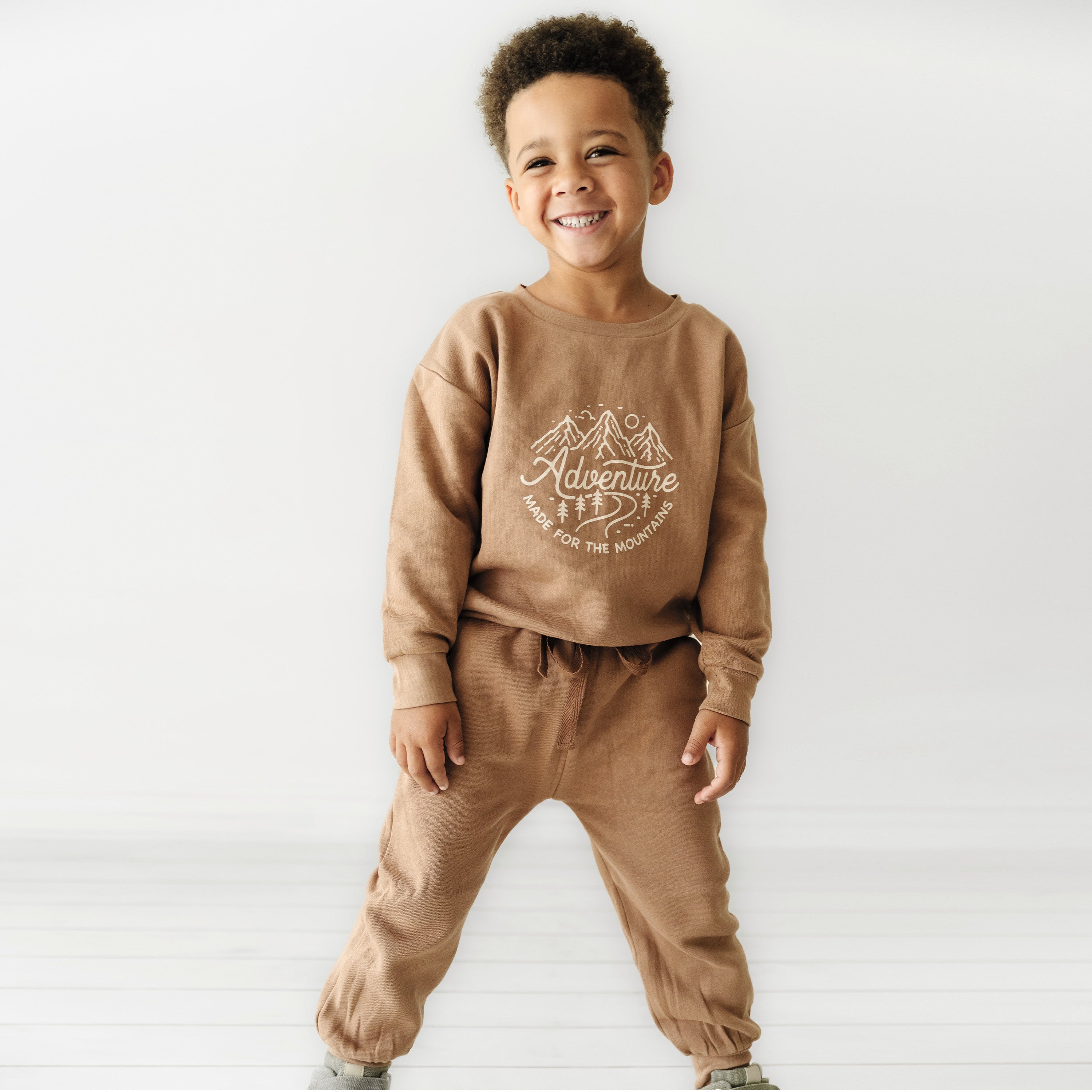 A young boy with curly hair smiles while wearing a brown sweatshirt and pants set from Organic Baby that reads "adventure" in white graphic text, standing confidently against a plain white background.