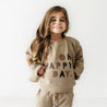 Young girl with long, wavy hair smiling, wearing a beige Organic Baby outfit with "oh happy day" printed on her shirt, standing against a white background.