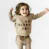 A joyful young girl in a beige Organic Baby outfit with the "Happy Day" Organic Graphic Sweatshirt printed on her sweatshirt is jumping in mid-air against a white background, smiling broadly with her arms out.