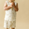 A toddler in a beige outfit with giraffe prints from Organic Kids holds a wooden stick, standing against a plain beige background. The child's face is not visible.