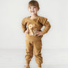 A young child in a tan Makemake Organics Organic Graphic Sweatshirt - Play and pants stands smiling in a brightly lit room with a plain white background.