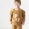A young girl with a ponytail smiles while wearing a mustard-colored sweatshirt and pants from Organic Baby with the word "play" written on the front, standing against a white background.