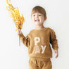 A joyful young girl with a ponytail, wearing an Organic Baby brown sweater with the word "play" on it, holding a bunch of yellow flowers, against a white background.