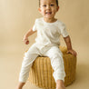 A joyful toddler in an Organic Kids Organic Tee & Pants Set with Wildcat Print sits on a wicker stool against a cream background, smiling broadly.