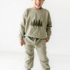 A young boy in an Organic Baby organic graphic sweatshirt and pants set with a forest design and the phrase "into the woods" stands smiling in a bright white room.