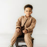 A young boy with curly hair sitting on a wooden stool, wearing a brown Organic Kids Sparkle quilted jacket and matching pants, smiling gently at the camera against a white background.