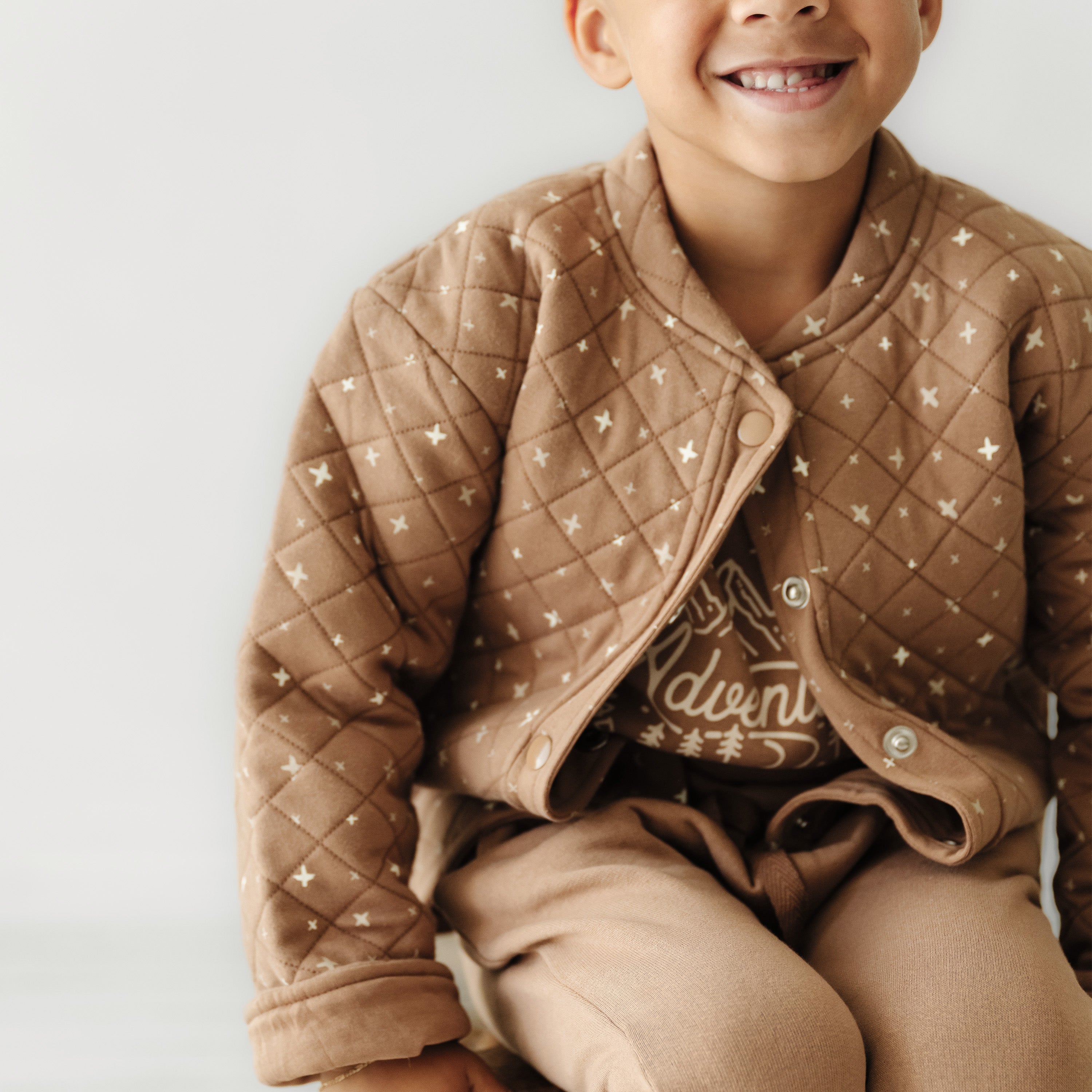 A young boy in a brown Organic Merino Wool Buttoned Jacket - Sparkle from Organic Kids, smiling joyfully against a light background. Only his upper body is visible, emphasizing his bright and cheerful demeanor