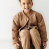 A young boy with curly hair smiling and sitting on a wooden stool wears Organic Kids Sparkle brown quilted jacket and matching pants. The background is plain and light-colored.