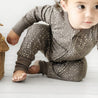 A baby in an Organic Baby speckled brown 2-Way Zip Romper crawls near a woven basket, looking to the side with curious eyes, in a bright, airy room.