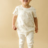 A young child in a white Organic Kids Wildcat Print outfit stands on a beige background, smiling with a chocolate in hand.