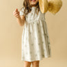 A young girl in a white Organic Kids Organic Puff Sleeve Dress - Tropical with a palm print, holding a straw hat, smiles playfully against a beige background.