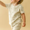 A joyful toddler in an Organic Kids Makemake Organics Organic Tee & Pants Set - Tropical outfit with a palm tree print is playfully running to the right, with arms outstretched and a blurred motion effect, on a warm beige.