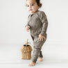 A toddler in an Organic Baby gray onesie with gold polka dots stands next to a small wicker basket on a white background, looking slightly to one side.
