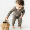 A toddler in an Organic Baby polka-dotted jumpsuit stands and reaches into a woven basket on a white background.