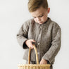 A young boy dressed in a Organic Kids' Organic Merino Wool Buttoned Jacket - Speckle looks into a woven basket he is holding, with a pensive expression. The background is plain and light-colored.