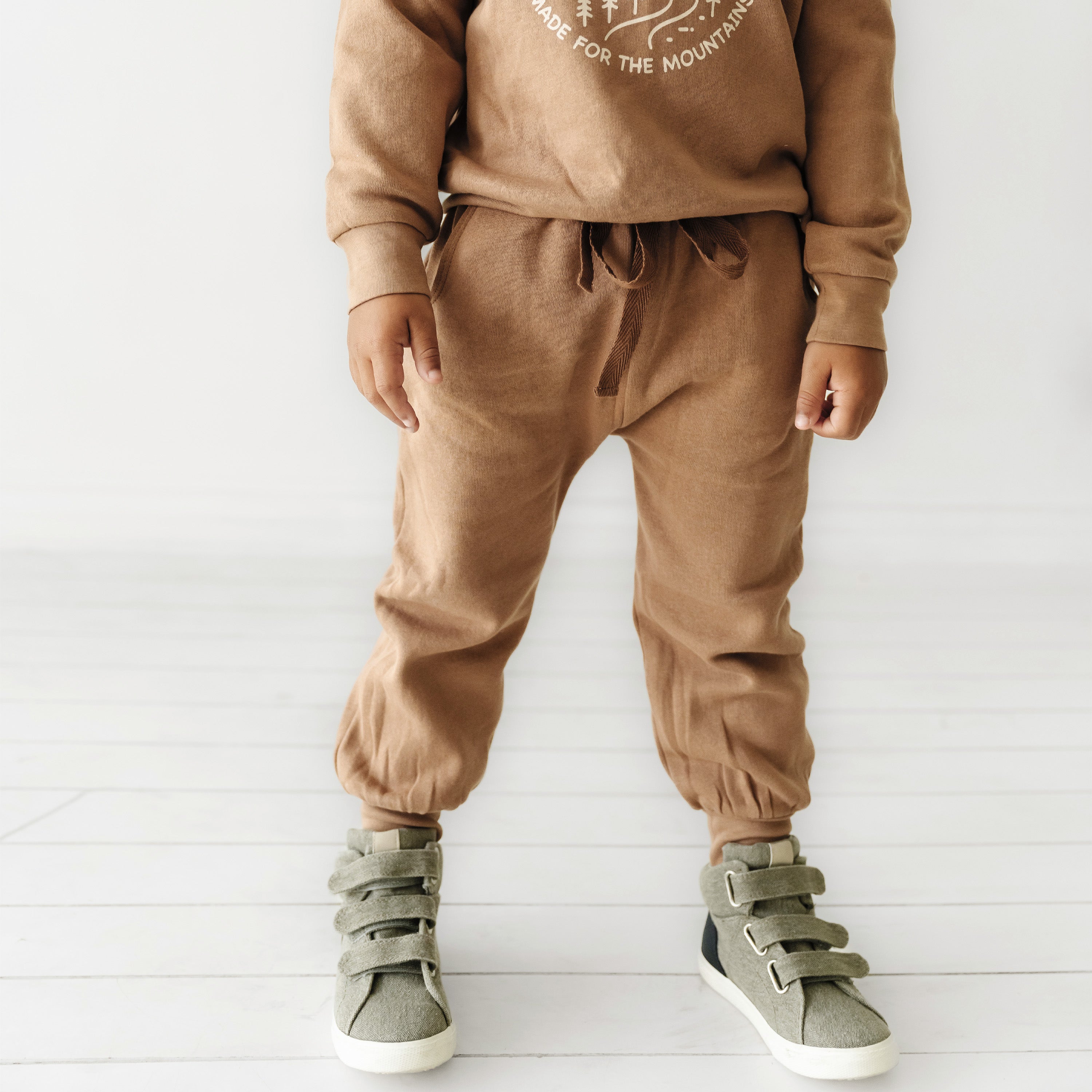 A toddler dressed in Organic Baby's Makemake Organics brown hoodie and matching sweatpants stands against a white background, showcasing green sneakers. Only the child's body from the neck down is visible.