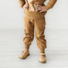 A child in an Organic Baby brown hoodie and Makemake Organics organic jogger pants outfit stands in a white room, only showing from the neck down. The outfit reads "play" and they wear light brown sandals.