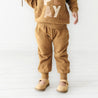 A young child wearing organic jogger pants in tan from Organic Baby with the word "play" on the hoodie, standing in a bright, minimalistic room.