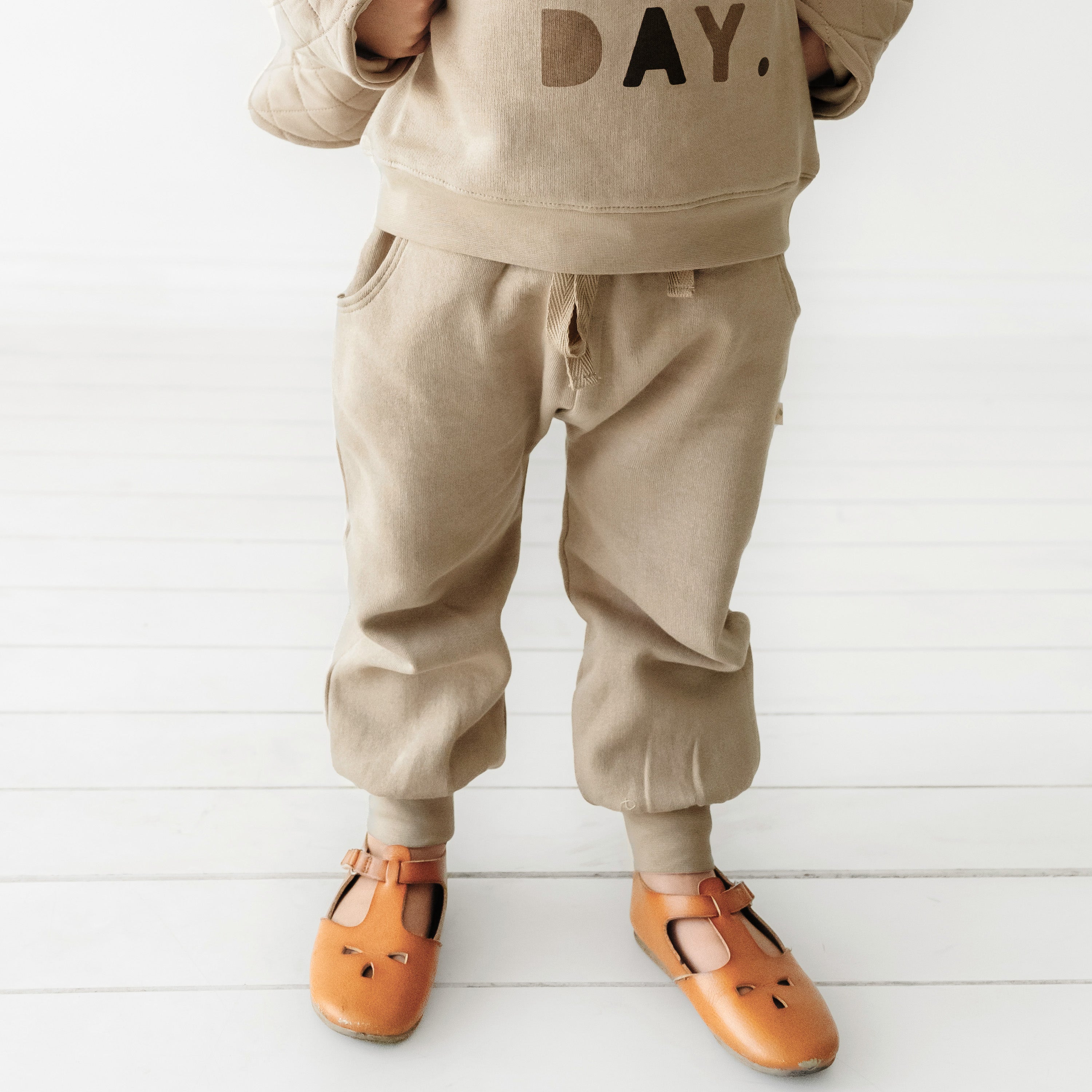 A child clad in Organic Baby's tan Organic Jogger Pants - Mocha and a matching sweatshirt labeled "day" from Makemake Organics stands on a white wooden floor, wearing orange fox-face sandals. Only the lower half of