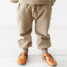 A child wearing Organic Baby Mocha organic jogger pants and orange animal face shoes stands against a white background, showing only from the waist down.