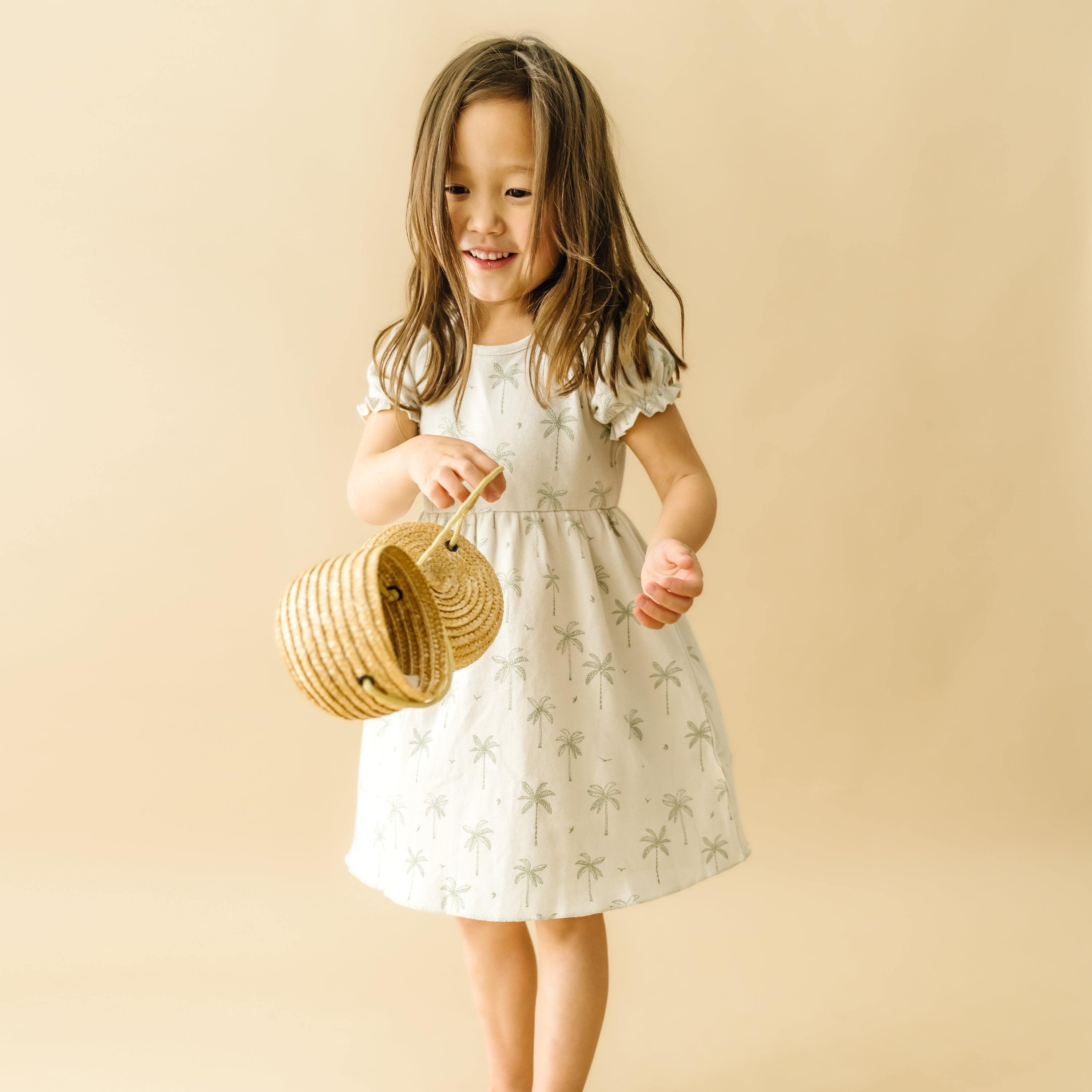 A joyful young girl in a white dress with Organic Kids palm prints, holding a wicker basket, playfully dancing against a soft beige background.