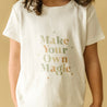 A child in a white Organic Tee & Pants Set from Organic Kids with the phrase "make your own magic" printed in decorative, gold and gray letters. The child's face is not visible.