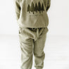 A child wearing olive Organic Baby jogger pants and a matching hoodie, standing facing away from the camera. The hoodie features a print of pine trees and the text "Into the Woods.
