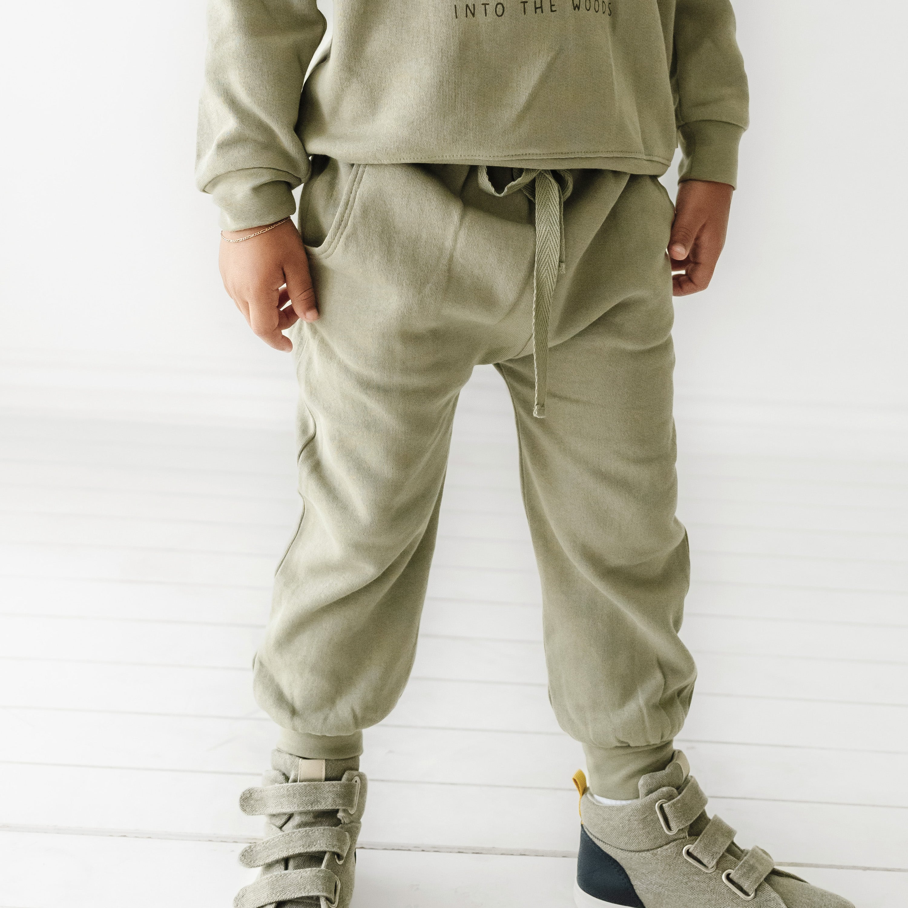 A child wearing Organic Baby's organic jogger pants in olive stands with hands in pockets, showing only the lower half of the body. The sweatshirt reads "into the