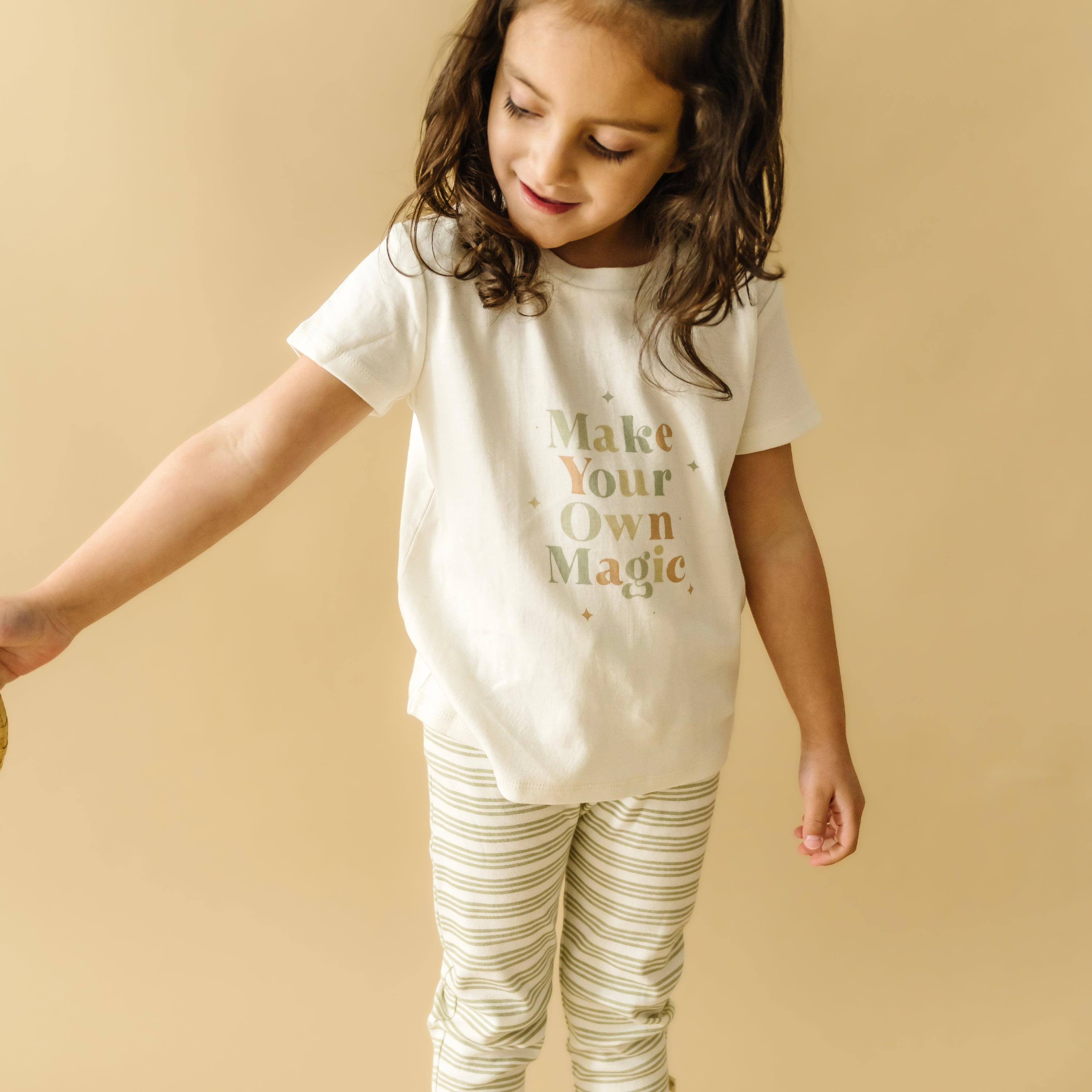 A young girl with dark hair wearing a white t-shirt from Organic Kids with the words "make your own magic" and striped pants looks downward with a content smile. She stands against a