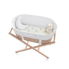 A modern Makemake Organics baby bassinet with a white quilted interior and beige wicker base, supported by a wooden frame. Inside is a small, plush teddy bear. The background is plain white.