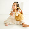 A toddler girl in a Makemake Organics Organic Linen Tiered Strap Dress - Beige Stripes sitting on the floor, holding a wicker basket filled with flowers, looking to the side with curiosity.