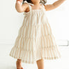 A toddler in a Organic Linen Tiered Strap Dress in Beige Stripes by Makemake Organics is toddling towards the camera in a brightly lit room, with visible excitement and motion. The focus is on her movement and the details of her dress.