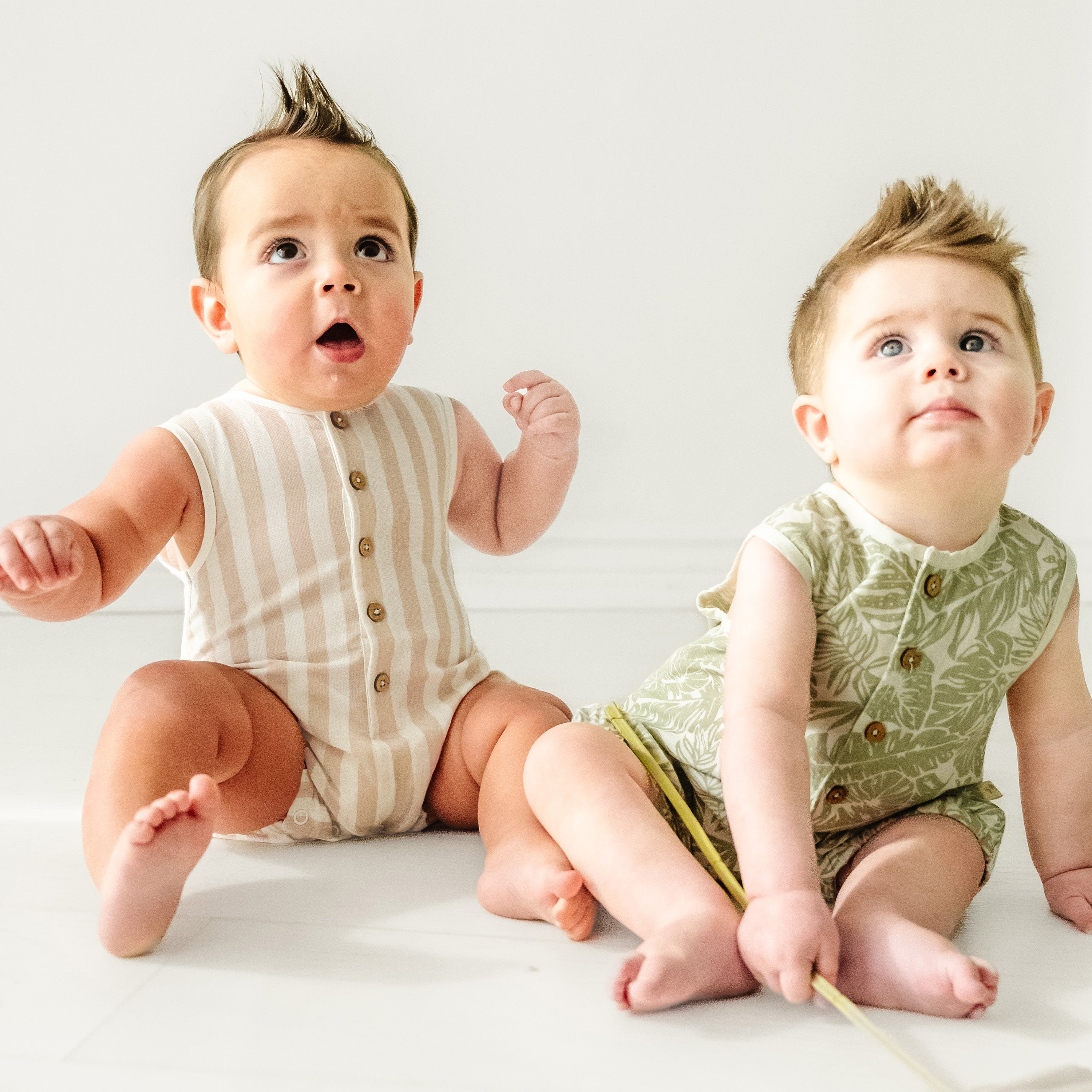 Two adorable baby girls with spiked hair sitting on a white floor, wearing stylish Makemake Organics Organic Sleeveless Bubble Romper in Beige Stripes. One looks surprised and the other holds a thin stick, both gazing towards something off-camera.