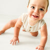 A cheerful toddler in a Organic Sleeveless Bubble Romper - Beige Stripes by Makemake Organics crawls towards the camera with an excited expression and bright eyes on a plain white background.