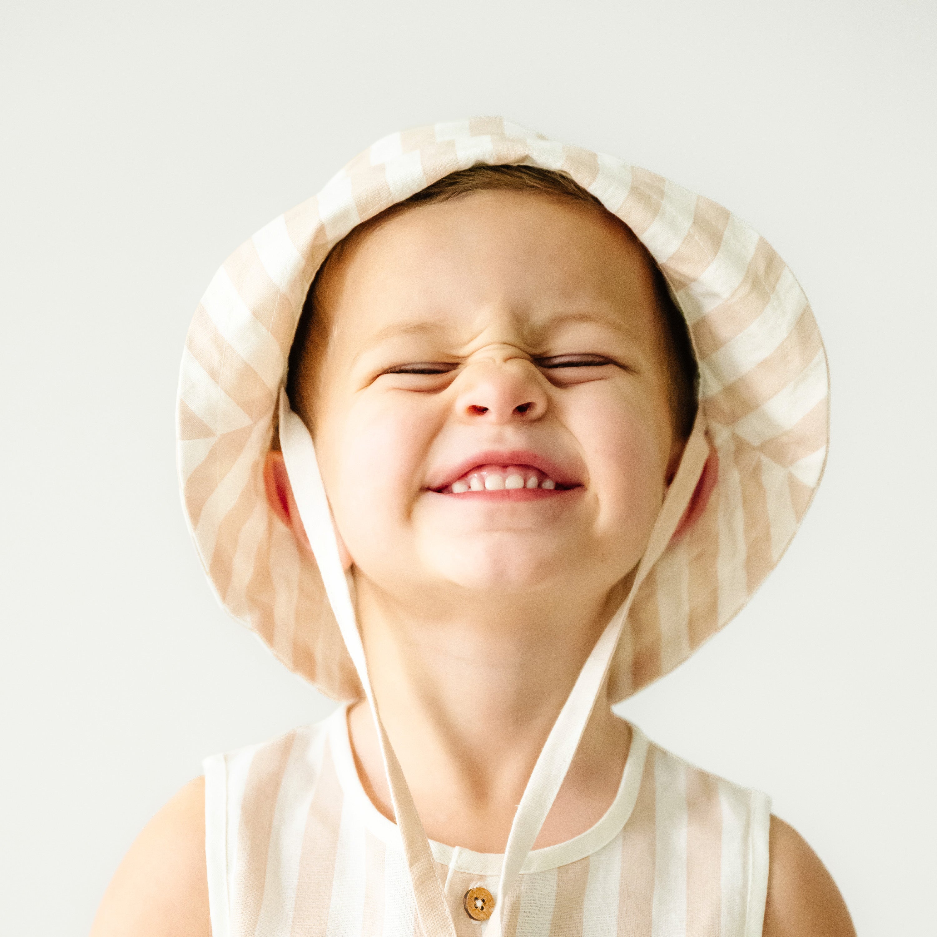 A cheerful young toddler with a wide, squinting smile, wearing a Makemake Organics Organic Linen Bucket Sun Hat in Beige Stripes and a sleeveless top against a plain background.