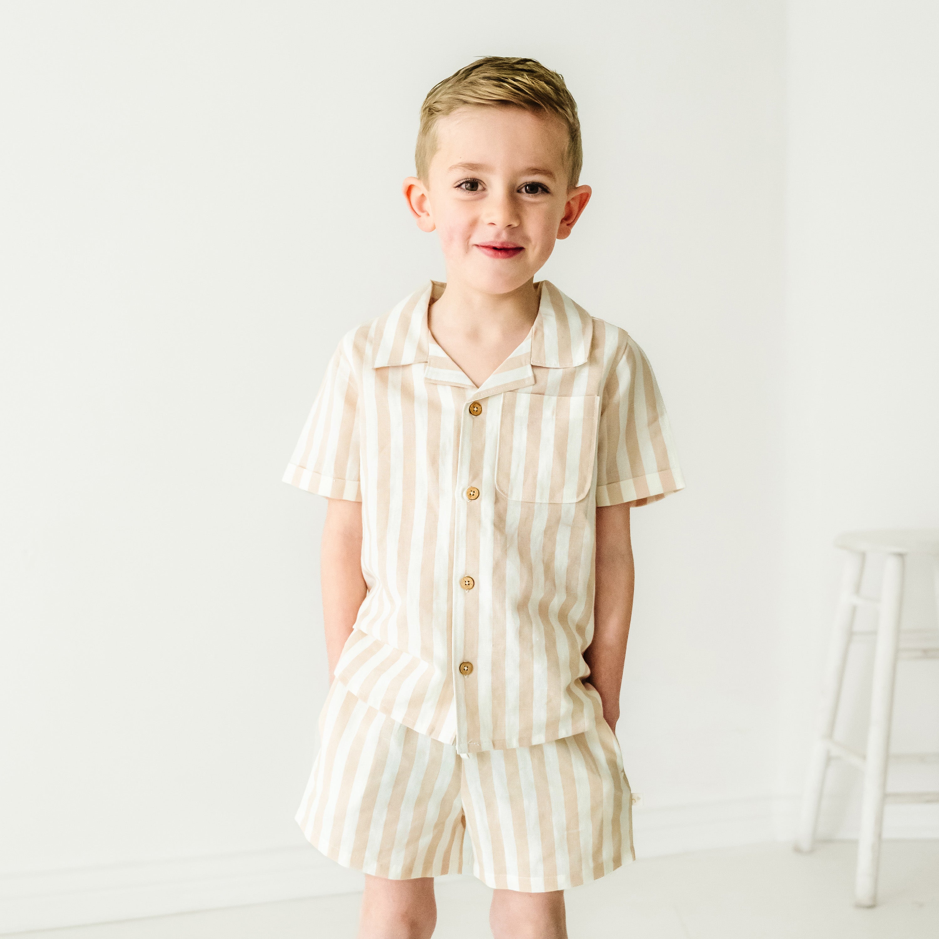 A toddler smiling at the camera, wearing a Makemake Organics Organic Linen Shirt and Shorts Set in Beige Stripes, standing in a bright white studio setting.