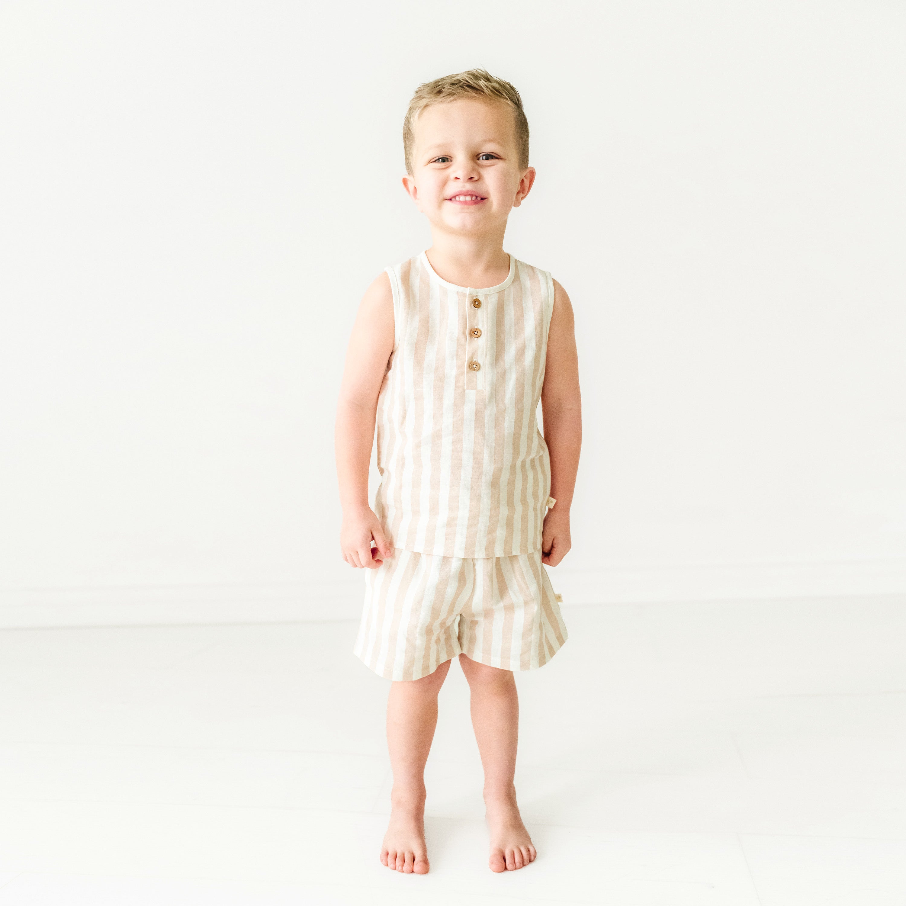 A young toddler smiles while standing barefoot, dressed in an Organic Linen Tank and Shorts Set in Beige Stripes by Makemake Organics in a bright, minimalist setting.