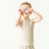 A young toddler in a Makemake Organics Organic Linen Tank and Shorts Set - Beige Stripes holds a large seashell over their eyes, standing against a plain white background.
