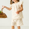 A young toddler with curly hair, wearing a Makemake Organics Organic Linen Flutter Top and Shorts in Beige Stripes and holding a wicker basket, stands in a light-colored room, pointing to something off-camera.